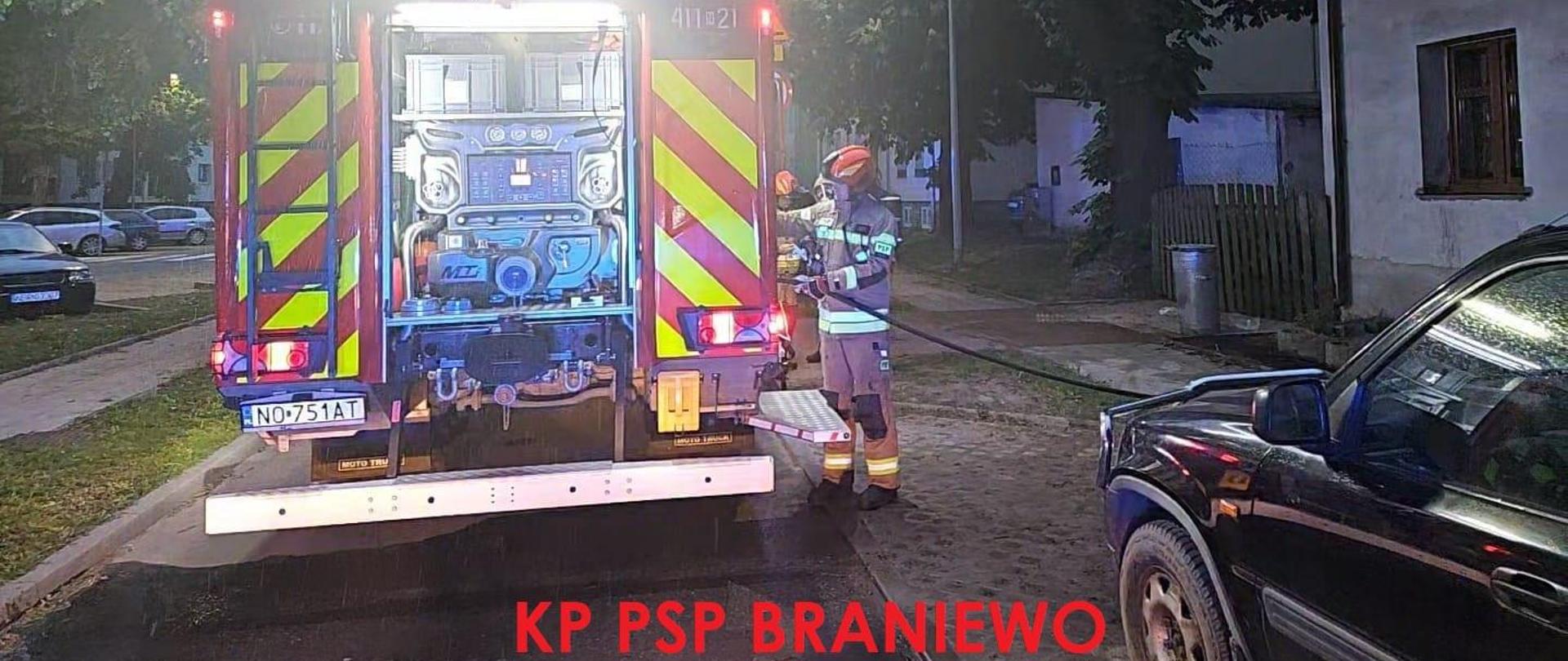 Na zdjęciu tył samochodu strażackiego. Przy samochodzie strażak. Z prawej strony budynek mieszkalny i samochód osobowy.