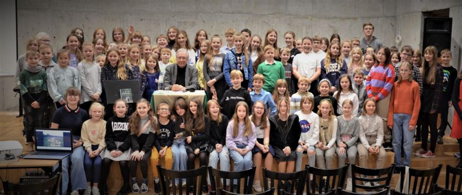 Zdjęcie przedstawia uczniów zgromadzonych na scenie wokół Krzysztofa Zanussiego