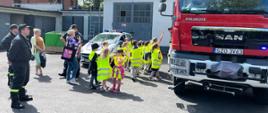 Dzieci w grupie stoją przy samochodach strażackim i policyjnym, obok nich opiekunki, strażacy, policjant. W tle budynek gospodarczy oraz drzew