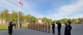 Funkcjonariusze Komendy Miejskiej Państwowej Straży Pożarnej w Elblągu stoją mundurach, na przeciwko siebie, w szeregu. W centralnej części znajduje się maszt z biało-czerwoną flagą. 