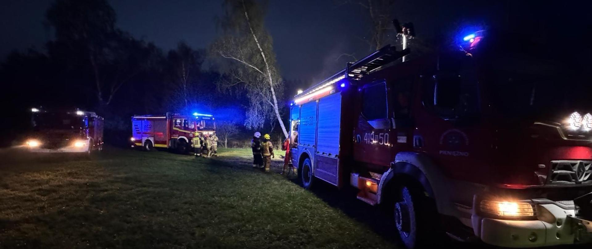 Zdjęcie zrobione nocą. Na zdjęciu stoją trzy samochody strażackie na światłach. Przy nich strażacy.