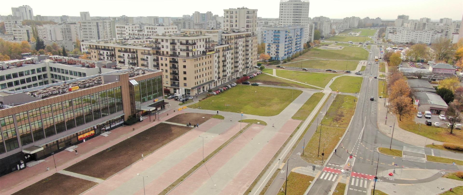 zdjęcie lotnicze terenu zabudowanego (pomiędzy blokami mieszkalnymi), pośrodku zdjęcia widoczny szeroki pas niezabudowany (jest to teren nad tunelem) i biegnącą wzdłuż niego ulicę. Ulica ta początkowo jest droga dwujezdniową, która przy pierwszej przecznicy zmienia się w jednojezdniową po jednym pasie w każdym kierunku. Wzdłuż ulicy ustawione są latarnie, po jej lewej stronie biegnie fragment ścieżki rowerowej również wyposażony w oświetlenie uliczne. Na dole fotografii widoczny jest duży parking wyłożony kostką.