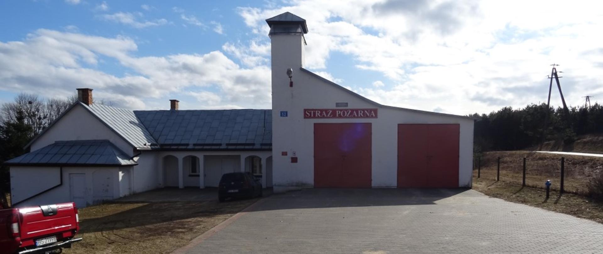 Widoczny jest budynek koloru białej elewacji z dwiema bramami koloru czerwonego. Na środku budynku usytuowana wieża. Dach z blachy koloru szarego.