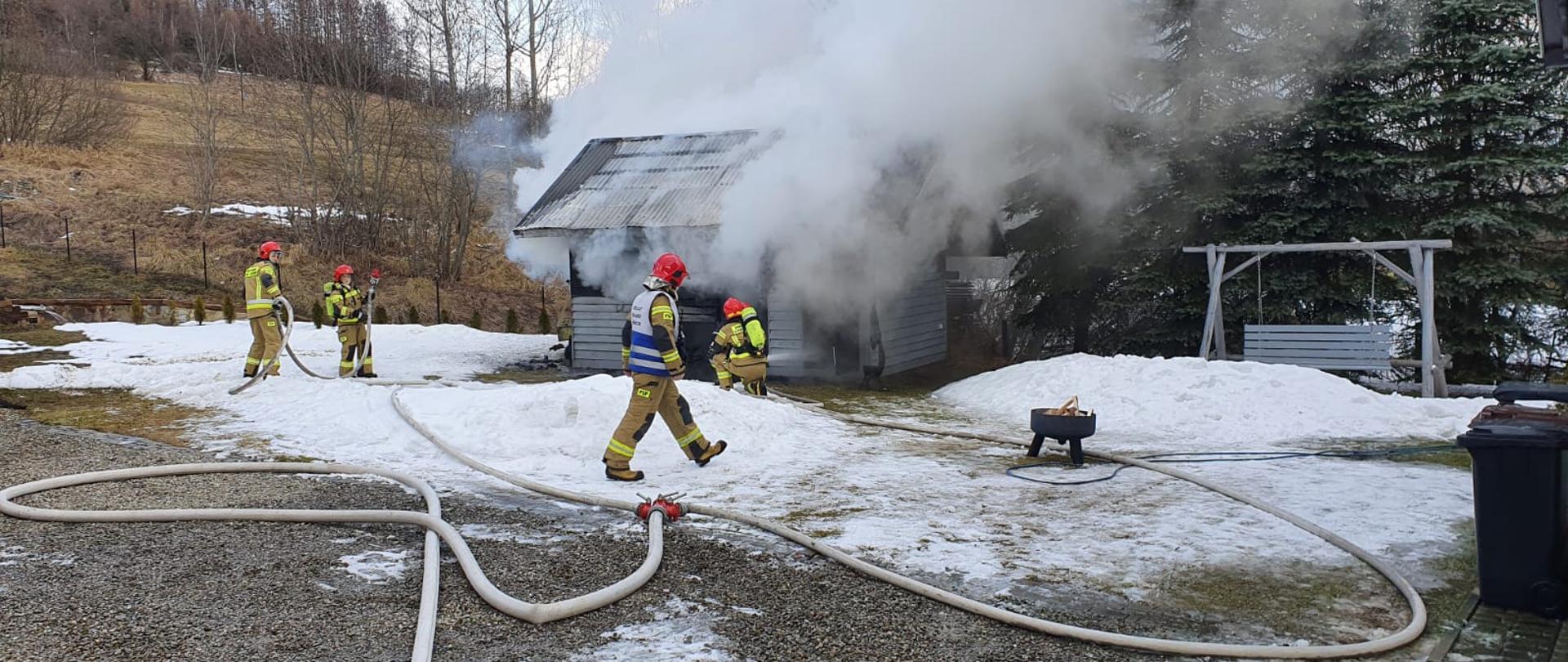Dym wydobywający się z płonącego budynku gospodarczego. Przed budynkiem strażacy. Na ziemi rozwinięta linia wężowa. 