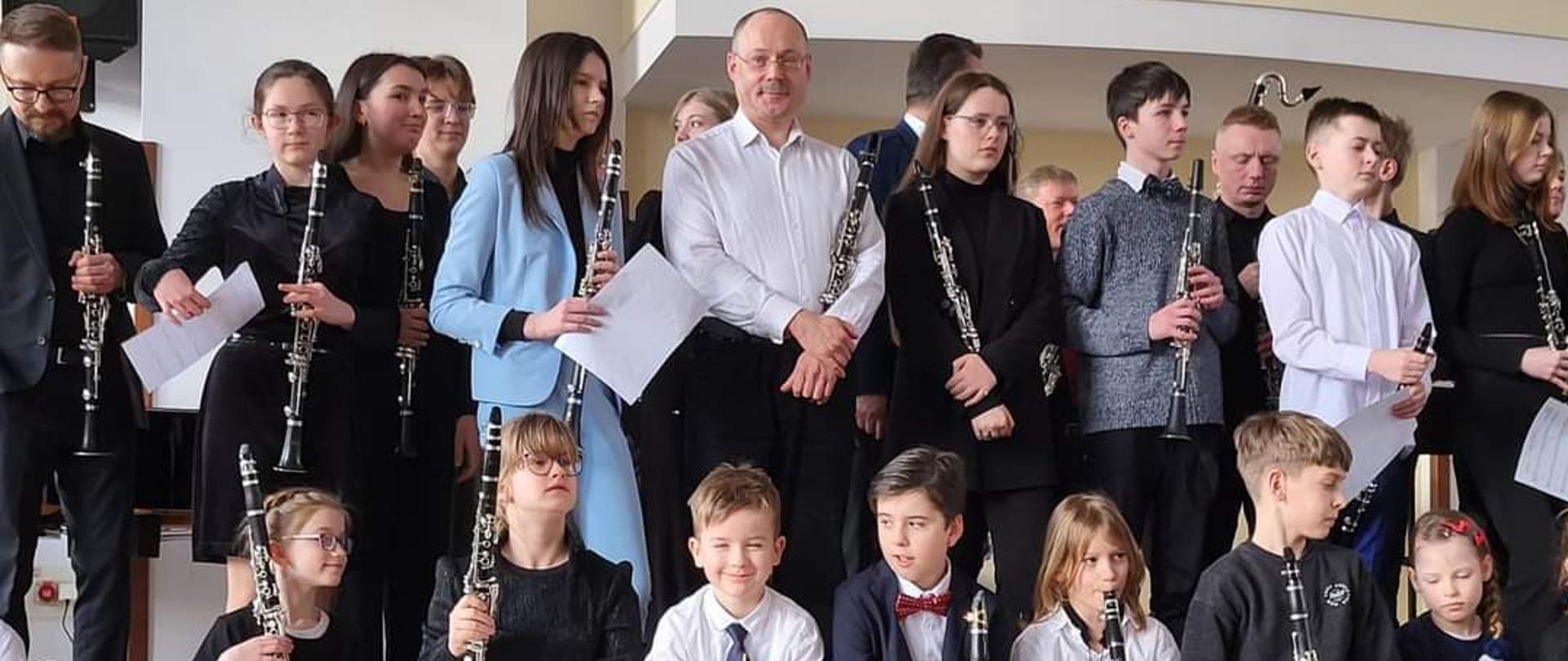 Fotografia przedstawia nauczyciela oraz grupę uczniów na scenie z klarnetami w dłoniach
