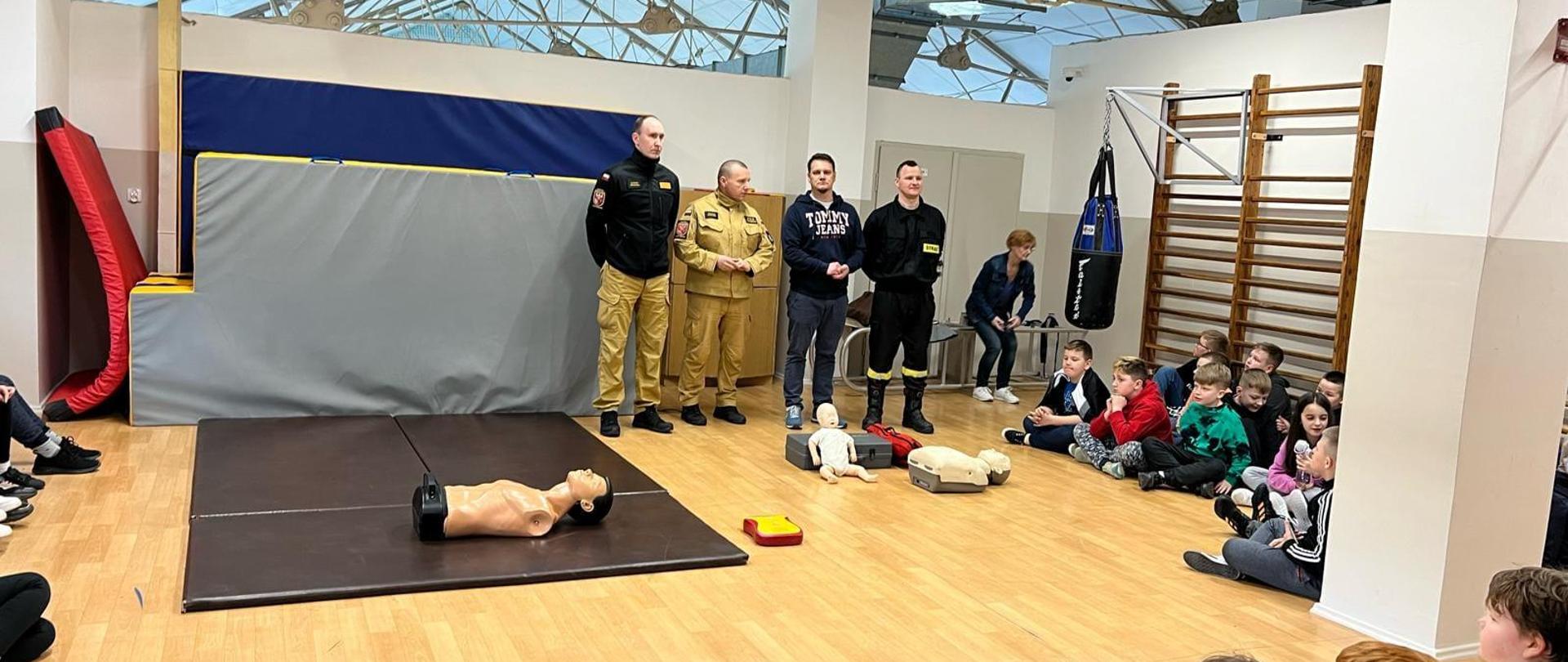 Strażacy na sali w okół siedzą dzieci, trwa szkolenie.