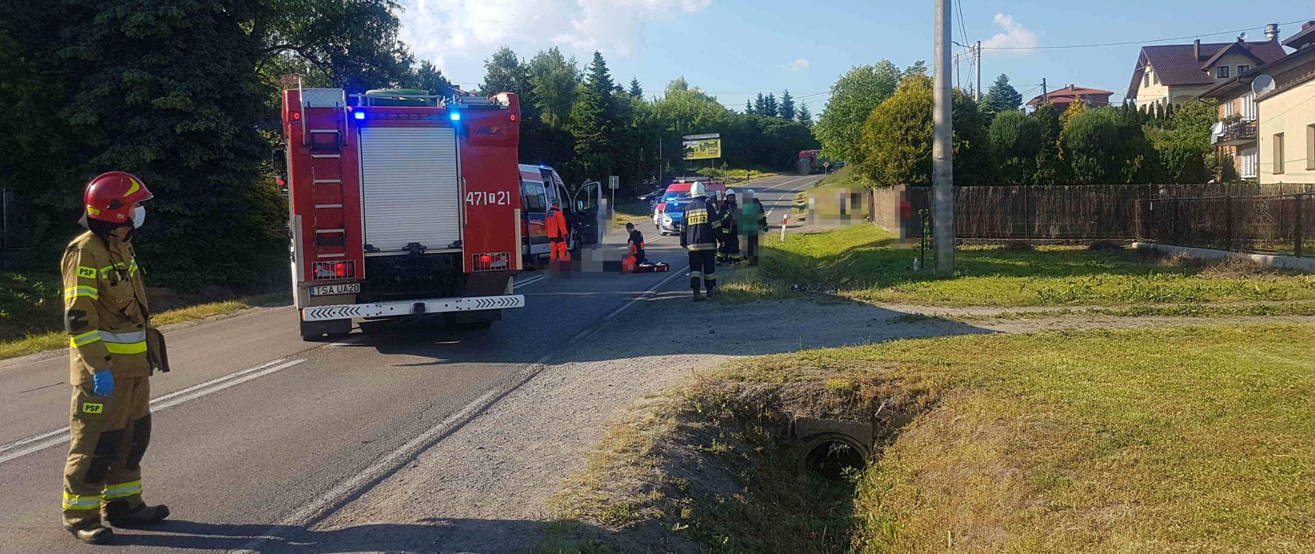 Zdjęcie przedstawia miejsce zdarzenia na pierwszym planie strażak zabezpiecza lądowisko LPR. Na dalszym planie widać samochód pożarniczy ambulans i ratowników medycznych udzielających pomocy poszkodowanemu.