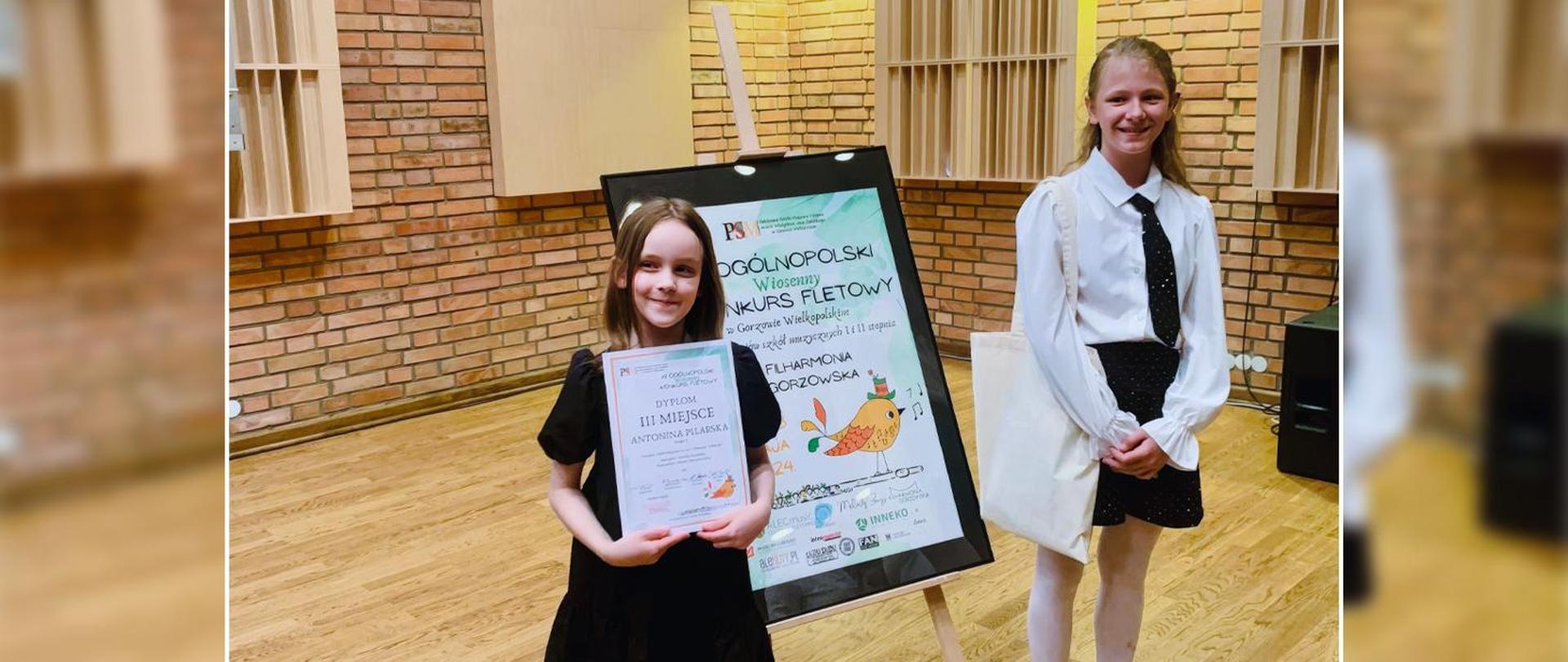Dwie dziewczynki stoją przy banerze reklamującym ogólnopolski konkurs fletowy, jedna z nich trzyma w ręku dyplom.