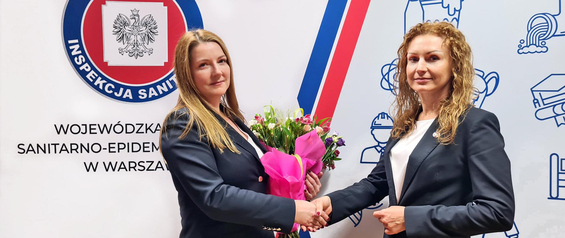 Dwie elegancko ubrane w garnitury kobiety podają sobie ręce. Jedna trzyma bukiet kwiatów. W tle logo Państwowej Inspekcji Sanitarnej.