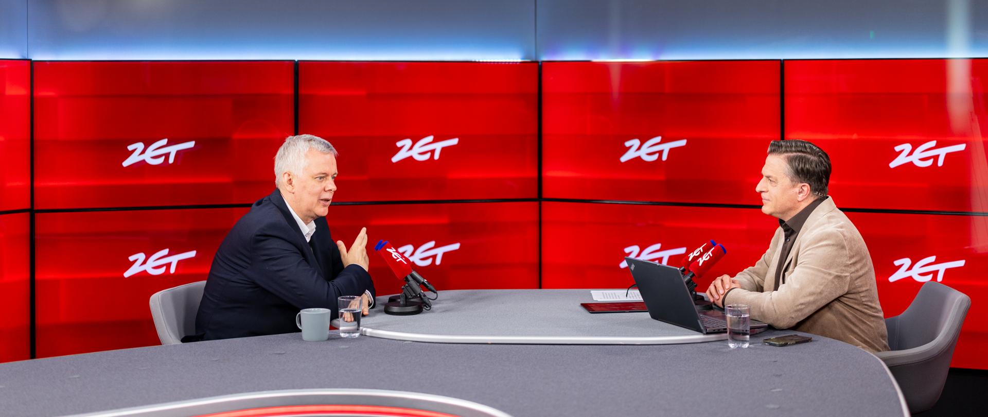 Na zdjęciu wnętrze studia radiowego. Przy szarym stole, na szarych krzesłach siedzą minister Tomasz Siemoniak (po lewej) oraz redaktor Bogdan Rymanowski (po prawej). Przed ministrem i redaktorem po dwa czerwone mikrofony z białym napisem "ZET". Przed redaktorem na stole - czarny laptop, szklanka z woda, telefon, kartki papieru, tablet. Przed ministrem szklanka z wodą oraz szary kubek. W tle ścianka wizyjna z wyświetlonymi białymi napisami "ZET" na czerwonym tle.