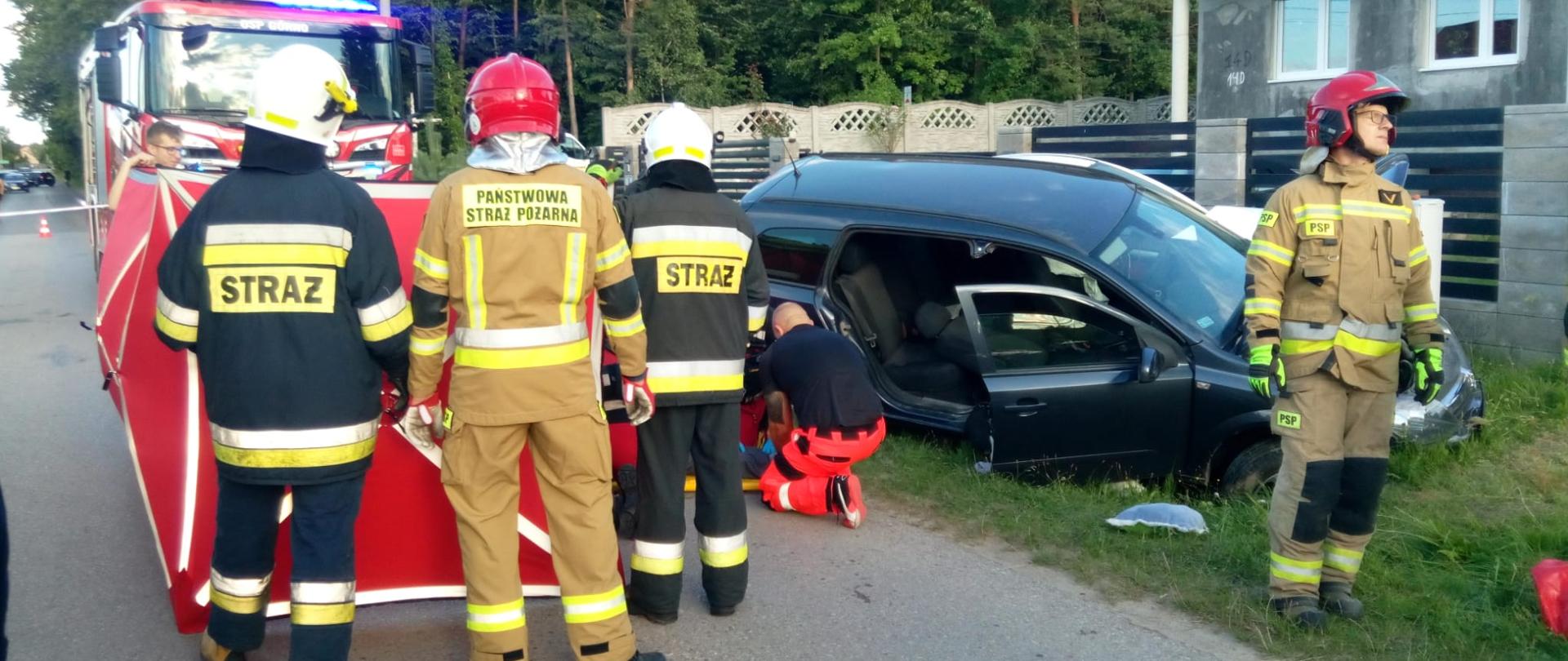 Zdjęcie przedstawia miejsce wypadku. Po prawej uszkodzony samochód, z lewej parawan osłaniający. Na pierwszym lanie ratownicy, a przy aucie klęczy ratownik medyczny. W głębi samochód pożarniczy.