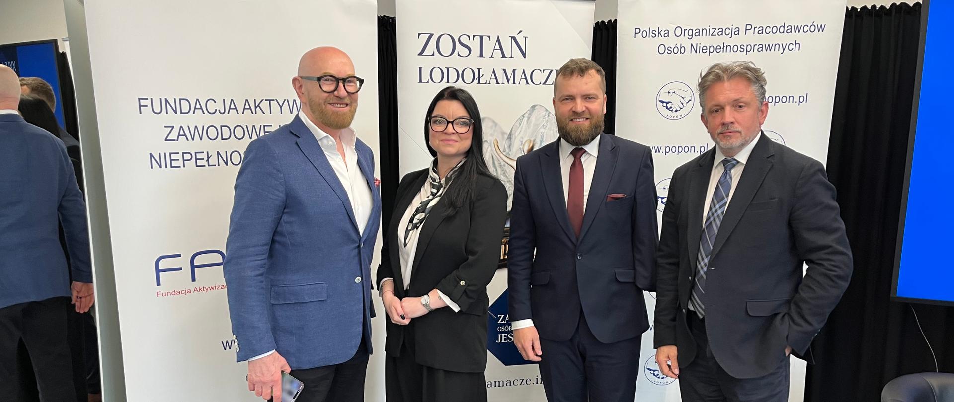 3 mężczyzn ubranych oficjalnie oraz kobieta w czarnym garniturze pozują na tle ścianek 