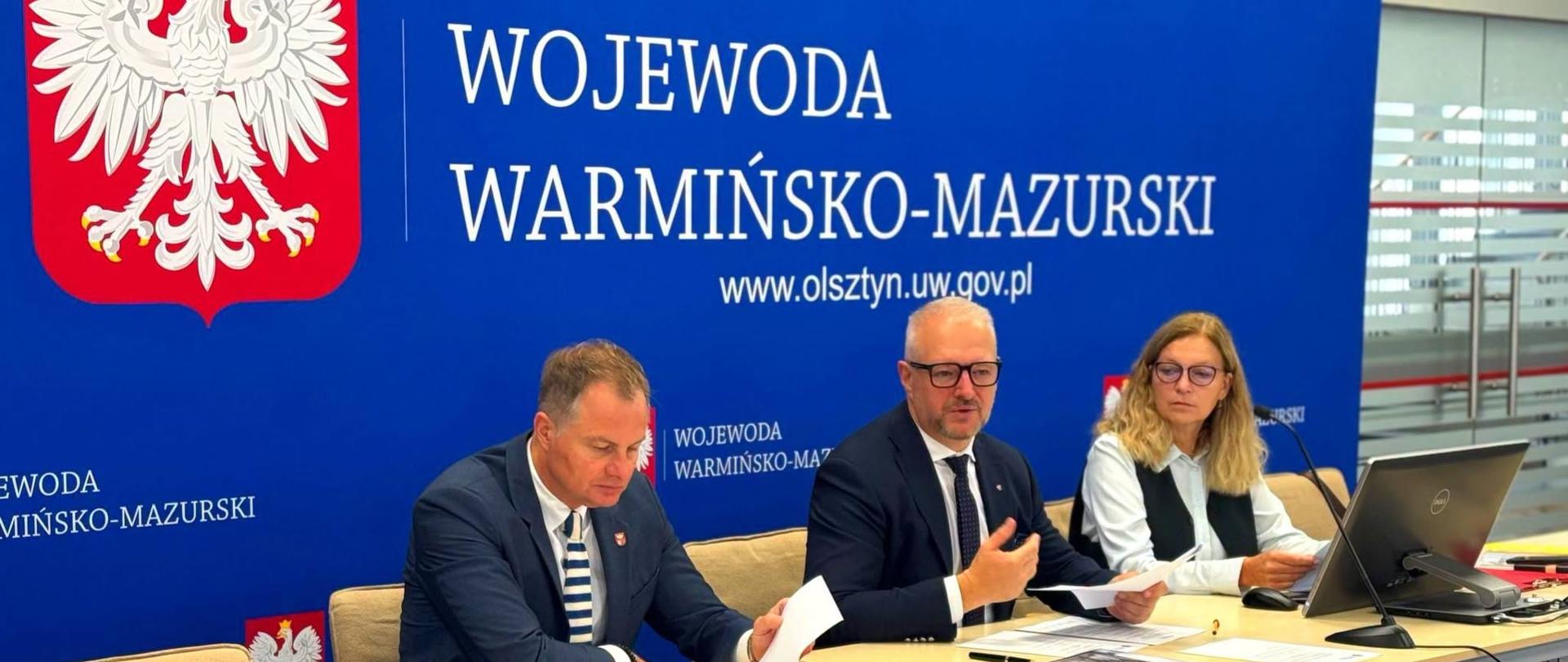 Trzy osoby, dwóch mężczyzn i kobieta, siedzą przy stole na tle dużej ścianki "Wojewoda Warmińsko-Mazurski". Wśród nich znajduje się wojewoda warmińsko-mazurski.