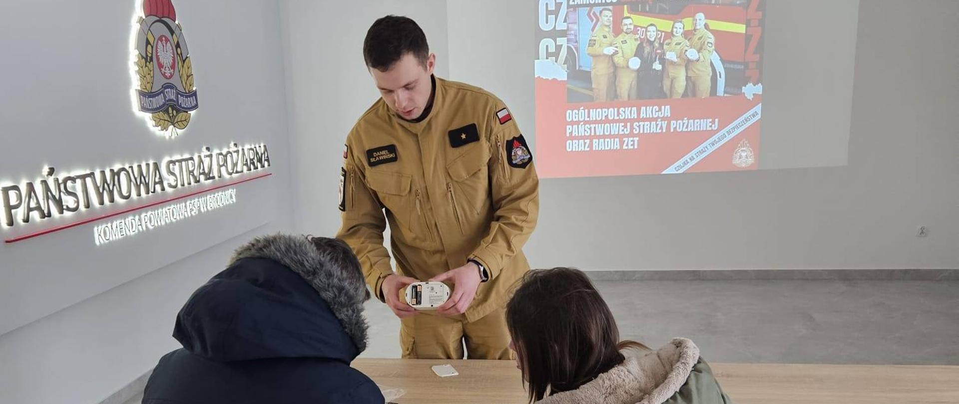 Zdjęcie przedstawia Funkcjonariusza PSP w Brodnicy prezentującego działanie czujki czadu Państwu, którzy zgłosili się po odbiór czujek dymu i czujek tlenku węgla – akcji „Czadowa Straż” prowadzonej wspólnie z Radiem Z. W tle wyświetlane jest logo akcji ze strażakami trzymającymi czujki. Po lewo na ścianie Logo i napis Państwowa Straż Pożarna.