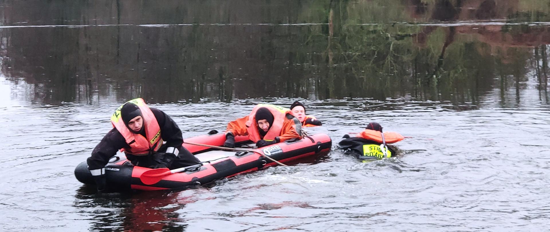 Na rozmarzającej tafli jeziora utrzymuje się w wodzie jedna osoba, w jej kierunku na desce lodowej płynie trzech ratowników w kamizelkazach z napisem STRAŻ na plecach.