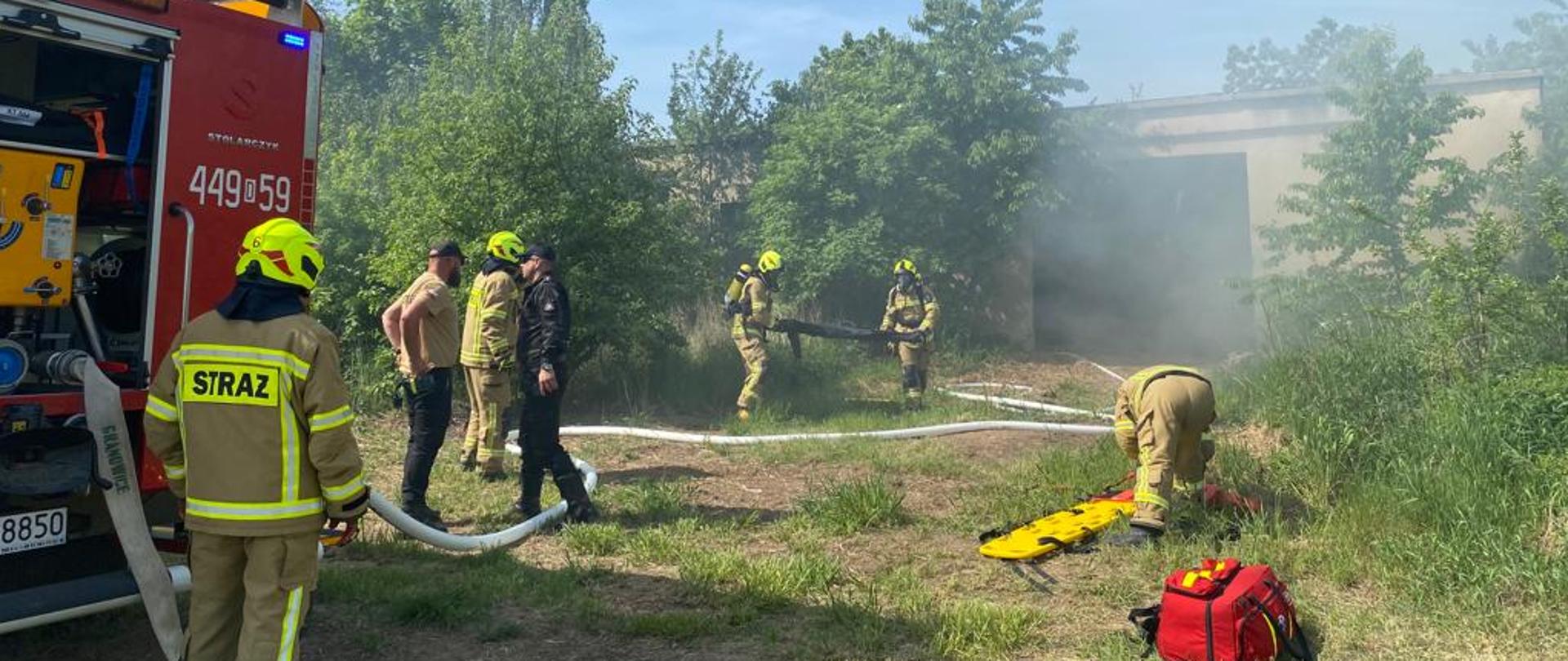 Obraz przedstawia strażaków w trakcie ćwiczeń praktycznych. Strażacy ewakuują osobę poszkodowaną na noszach. Widać rozwinięte linie gaśnicze. Strażacy w umundurowaniu specjalnym. W tle zadymiony budynek. Pora dzienna.