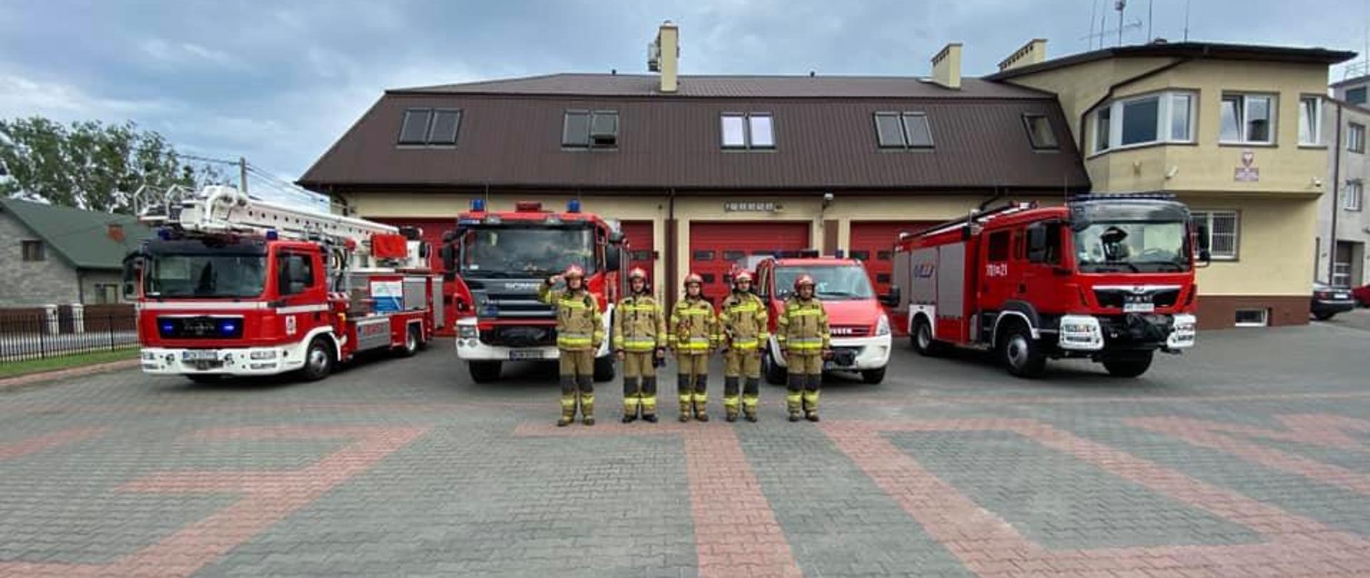 Strażacy z Komendy Powiatowej PSP w Zwoleniu oddają hołd Powstańcom w 77. rocznicę wybuchu Powstania Warszawskiego. Stoją w ubraniach specjalnych przed strażnicą oaz przed samochodami bojowymi.