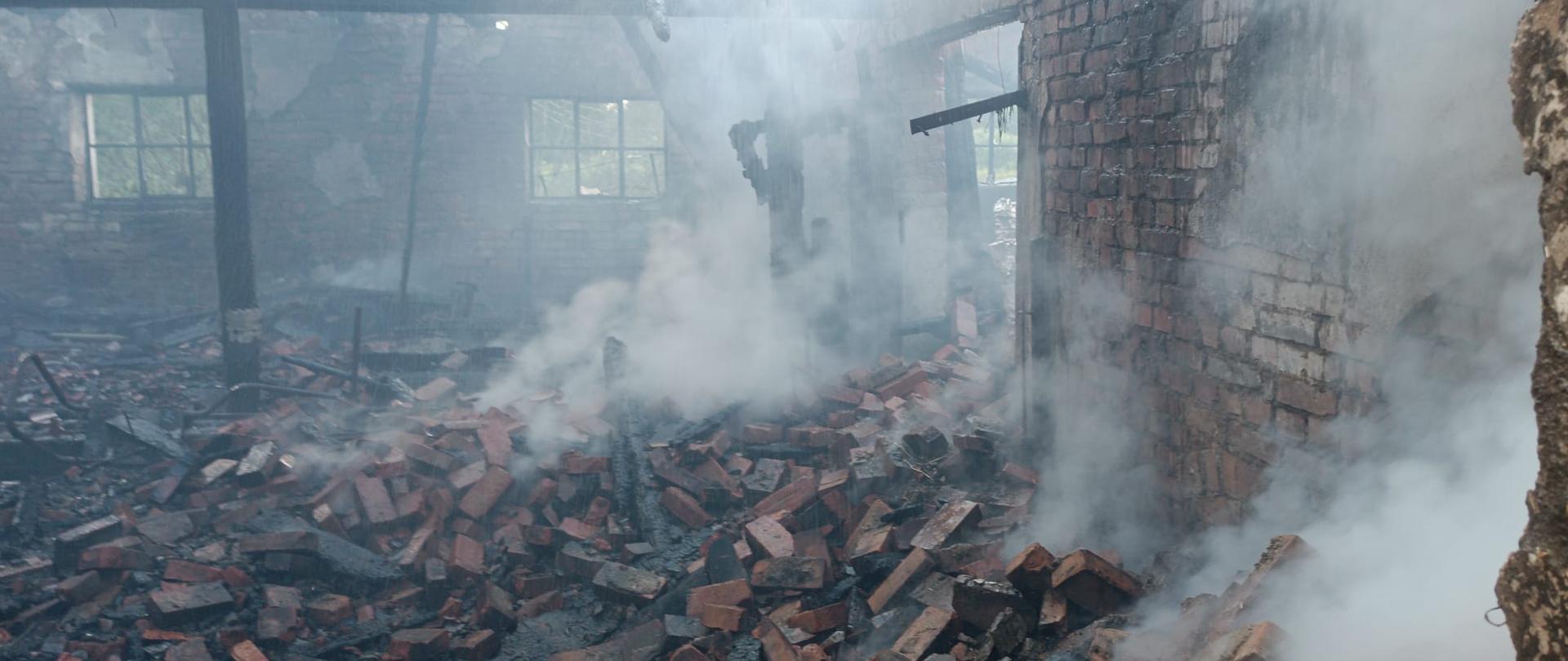 Spalone wnętrze budynku gospodarczego. Nadpalona drewniana konstrukcja, osmalone belki i elementy więźby dachowej. na pierwszym planie widoczne cegły z zawalonej, murowanej części obiektu. Nad pogorzeliskiem unosi się duża ilość dymu.