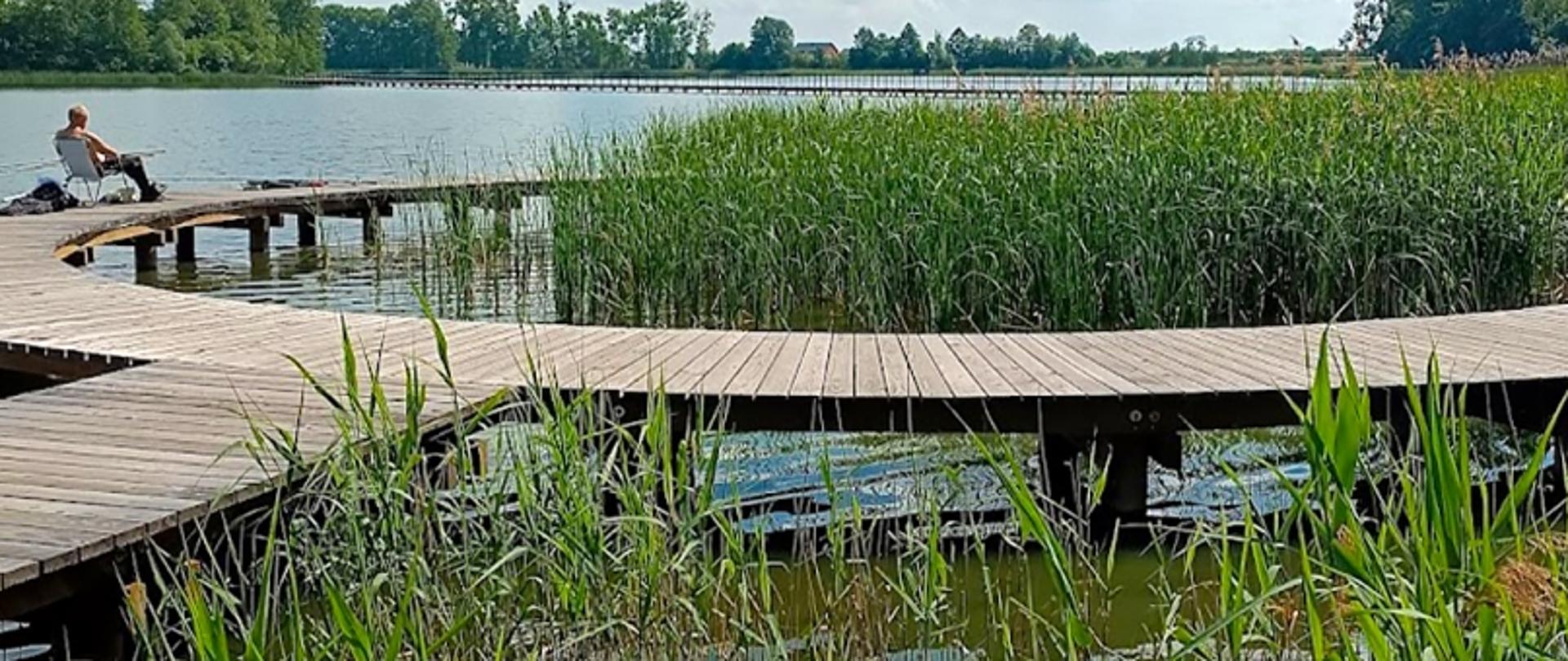 Osoba z wędką siedząca na pomoście nad jeziorem, w otoczeniu traw i drzew.