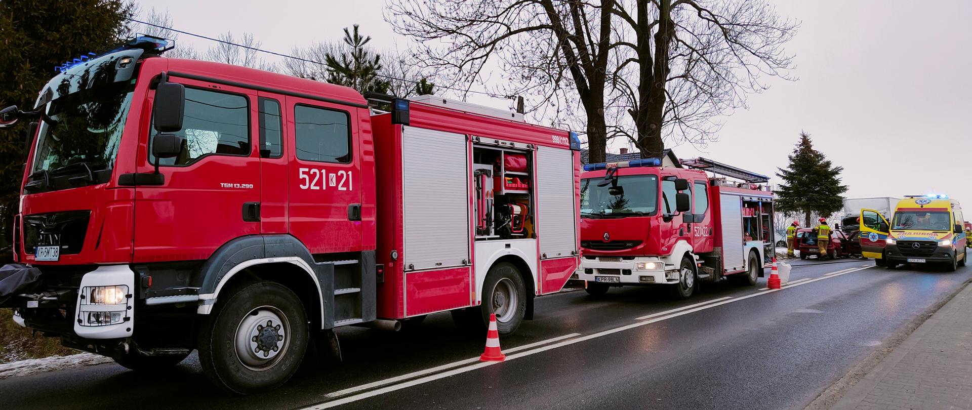 Wozy strażackie na pierwszym planie a w tle rozbity samochód i karetka pogotowia