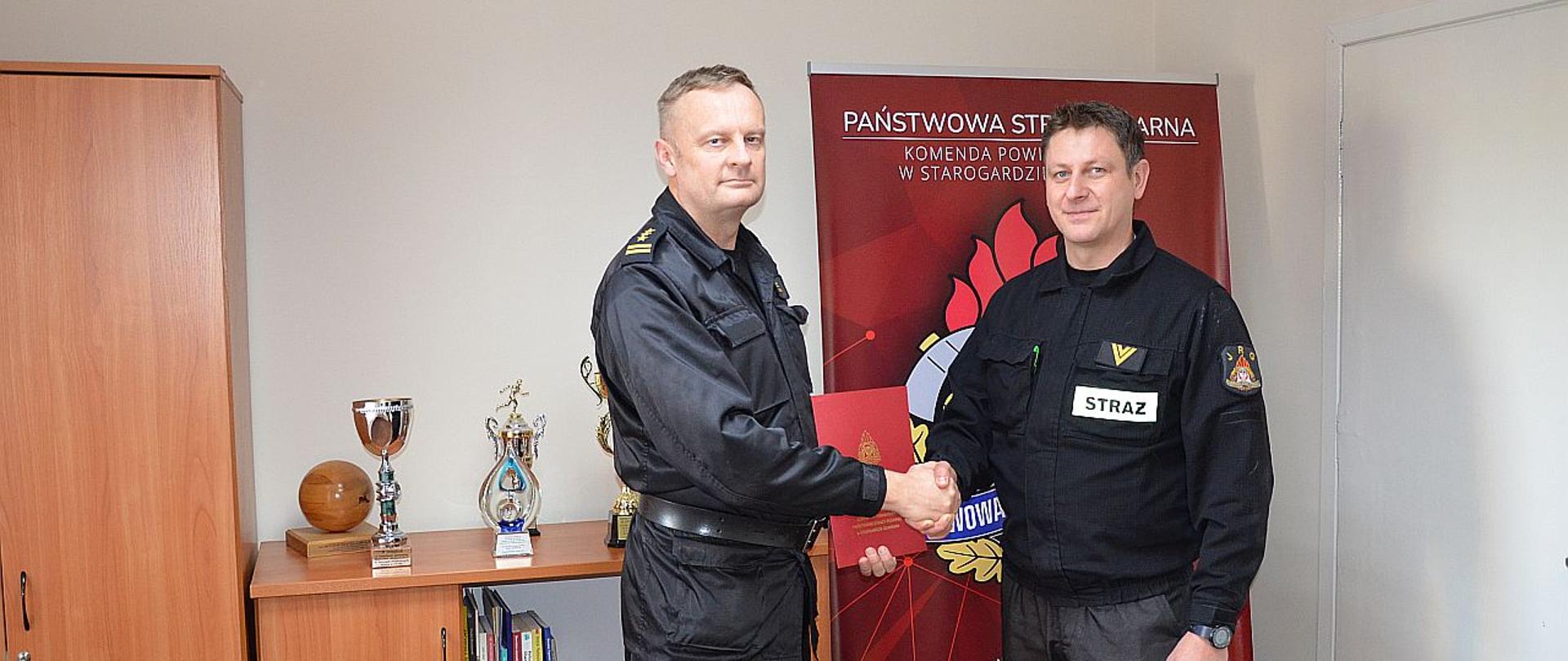 Zdjęcie przedstawia moment gratulacji. W tle baner Komendy, puchary i nagrody. Komendant powiatowy gratuluje uściskiem dłoni wyróżnionemu strażakowi.