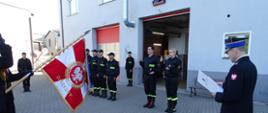 Zdjęcie wykonano w ciągu dnia na placu Komendy Powiatowej Państwowej Straży Pożarnej w Sępólnie Krajeńskim. Na pierwszym planie Komendant Powiatowy Państwowej Straży Pożarnej w Sępólnie Krajeńskim w mundurze galowym. Z lewej strony poczet sztandarowy z opuszczonym sztandarem, naprzeciw dwoje strażaków w mundurach koszarowych podczas ślubowania. W tle zmiana służbowa w mundurach koszarowych. 