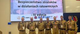 Grupa siedmiu strażaków w piaskowych mundurach służbowych stoi w jednym rzędzie na scenie sali konferencyjnej Akademii Pożarniczej. Za nimi znajduje się wielki ekran z oficjalnym tłem wydarzenia oraz napisem: „Konferencja naukowa, 20-21 października 2026 r., Bezpieczeństwo strażaków w działaniach ratowniczych – IV edycja” wraz z logotypami organizatorów i patronów. 
