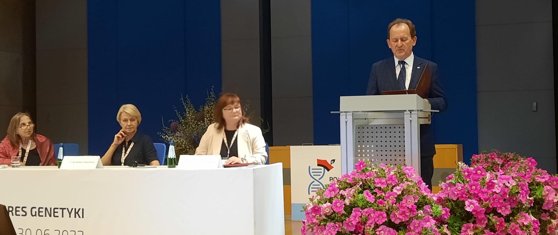 Minister Bernacki stoi na mównicy i mówi do mikrofonu, obok bukiety różowych kwiatów i podwyższenie, przy którym siedzą trzy kobiety.