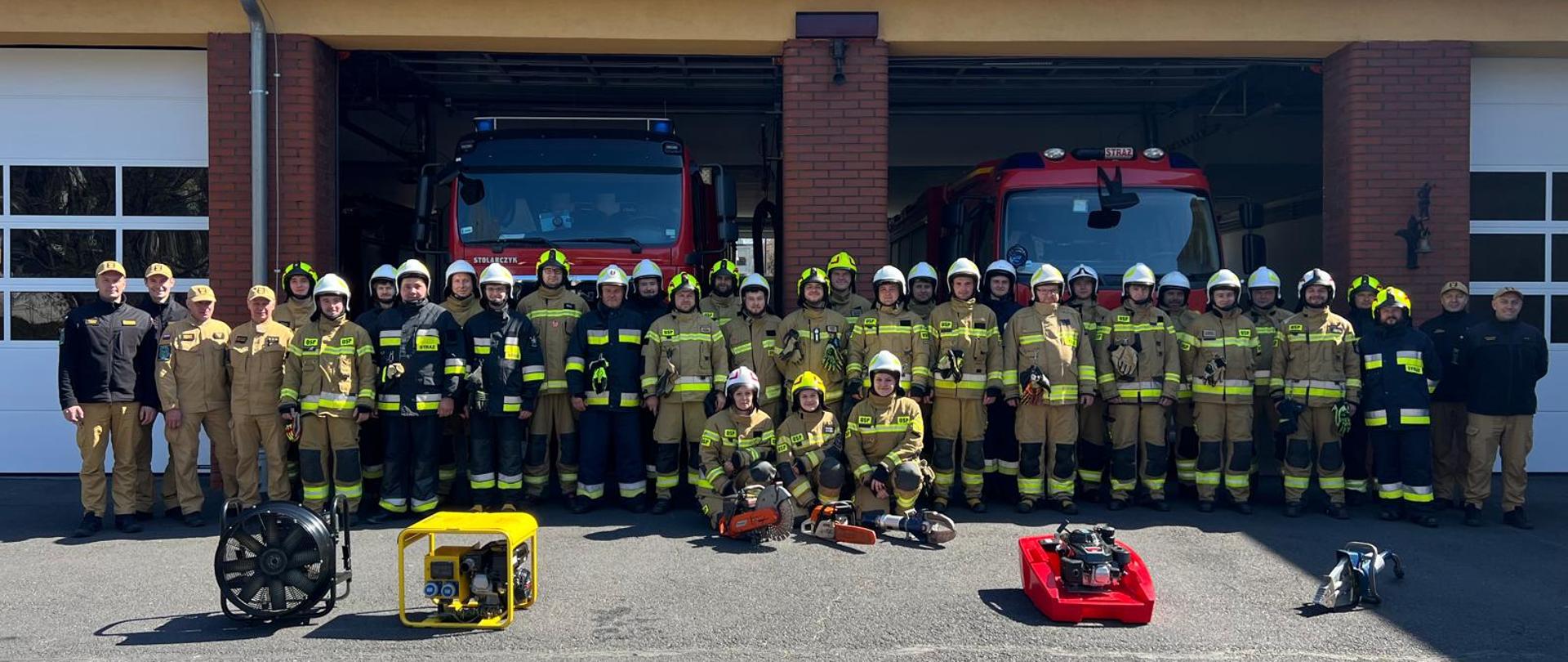 Na tle budynku i samochodów pożarniczych JRG Olesno ustawieni są uczestnicy kursu wraz z komisją egzaminacyjną i instruktorami. Przed strażakami ustawiony jest sprzęt ratowniczy.