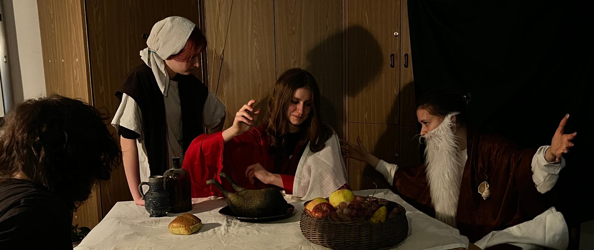 Zdjęcie przedstawia inscenizowaną scenę z obrazu "Uczta w Emaus" Caravaggia, odegraną w pomieszczeniu szkolnym.
Kilka postaci siedzących i stojących wokół stołu przykrytego białym obrusem oraz brązowym papierem.
Stół jest ustawiony na pierwszym planie. Na nim znajdują się:
kosz z owocami (jabłka, gruszki),
duży dzban,
bochenek pieczywa,
półmisek z przygotowaną potrawą.
Uczestnicy mają na sobie kostiumy stylizowane na dawne stroje, m.in. długie tuniki, płaszcze, nakrycia głowy, sztuczną brodę.
Oświetlenie jest przyciemnione, kierunkowe, co nadaje scenie teatralny charakter.
W tle stoją drewniane szafki, a po prawej stronie znajduje się ciemne materiałowe tło.