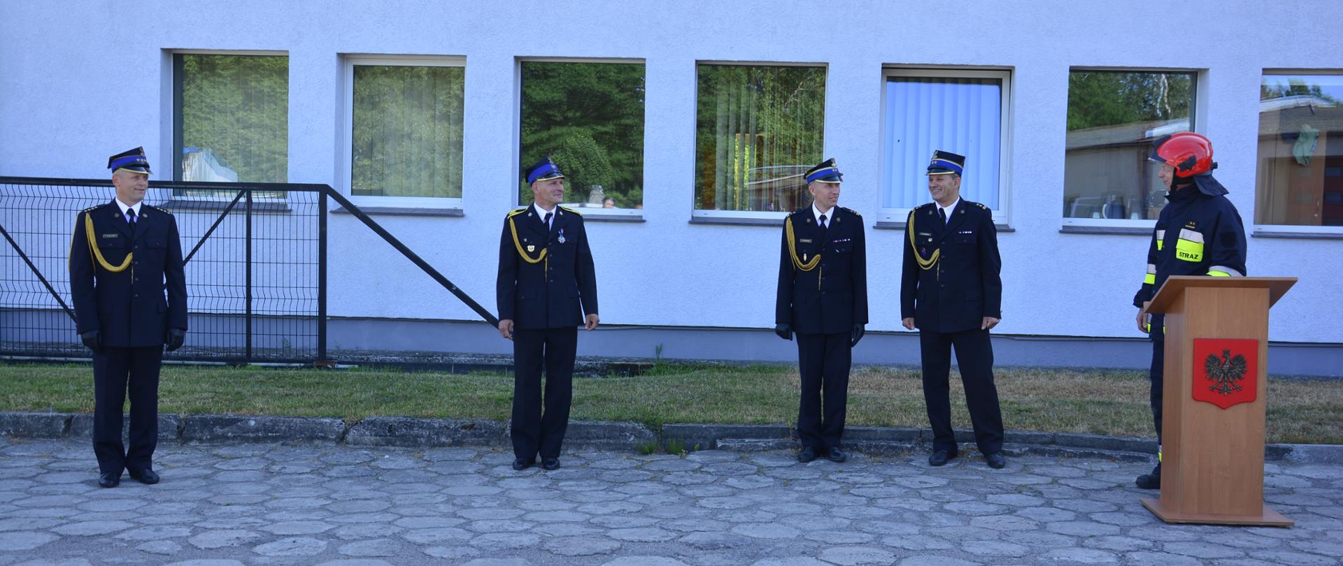 Zdjęcie przedstawia czterech strażaków ubranych w mundury galowe przed budynkiem. Piaty ubrany u ubranie specjalne oraz hełm stoi przy mównicy.