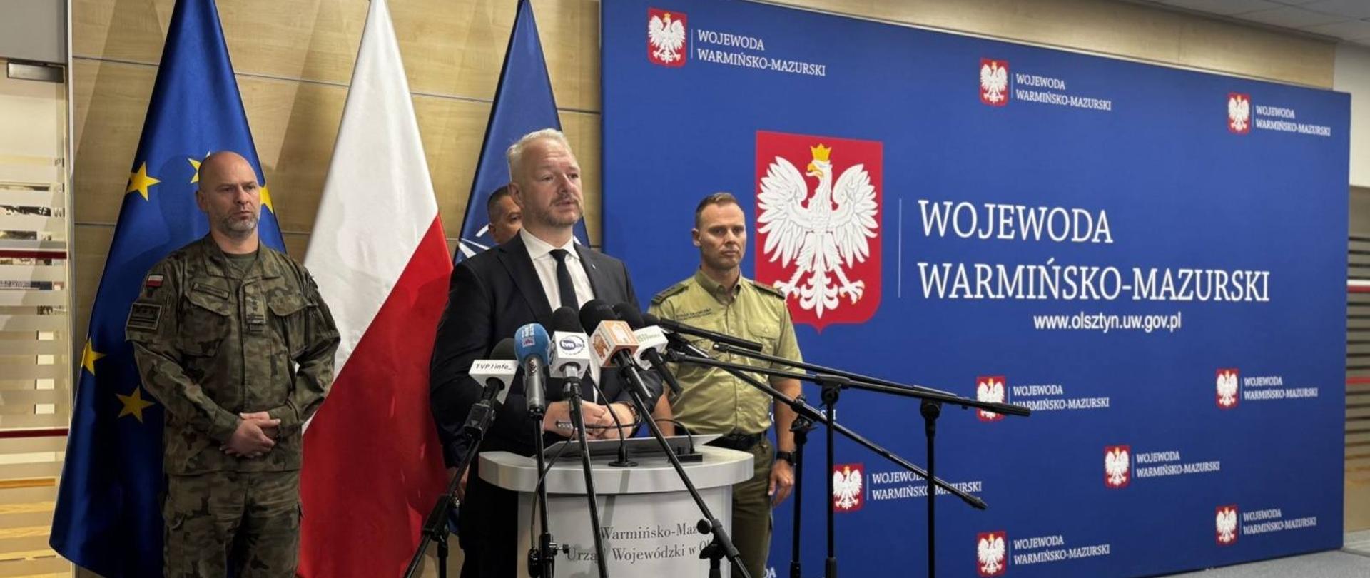 Mężczyzna w sali z flagami przemawia przez mikrofon, za nim stoi trzech mężczyzn w mundurach. 