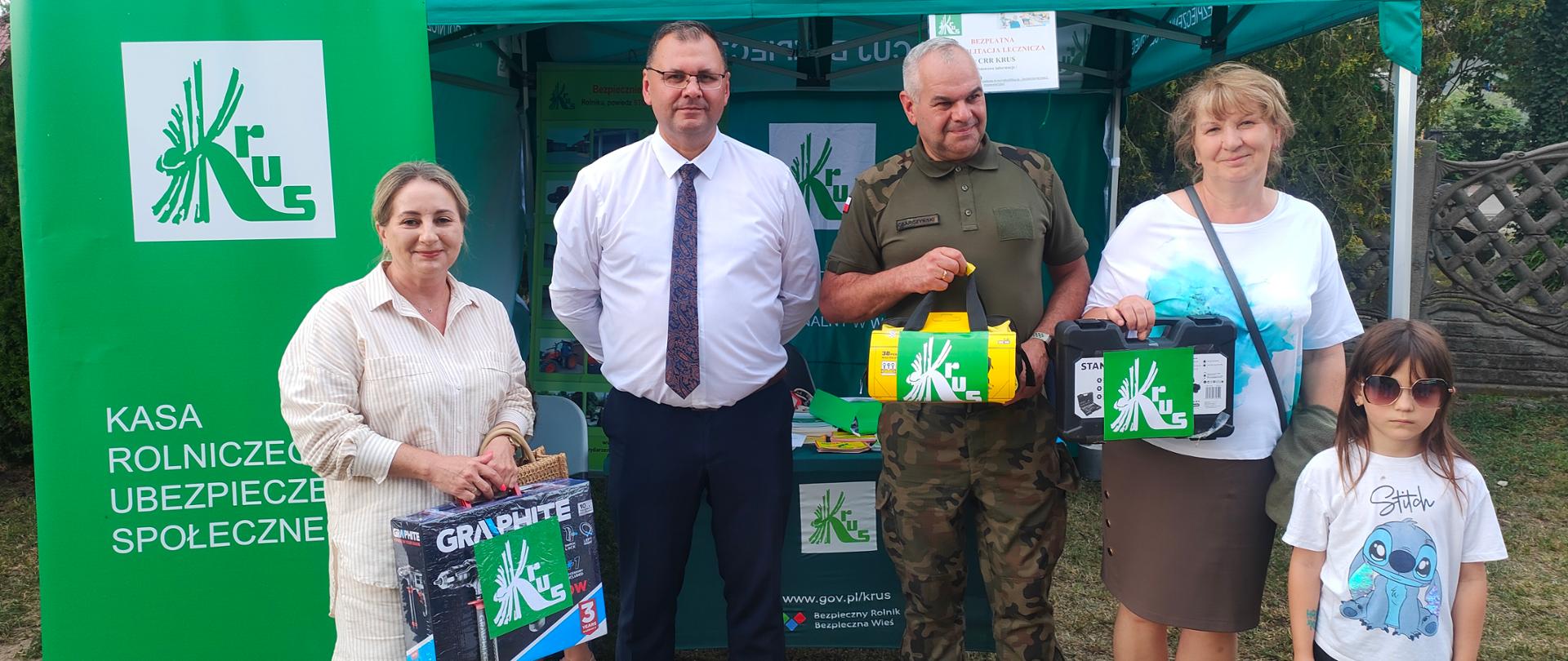 Mężczyzna w białej koszuli i w krawacie stoi przy stoisku Kasy razem z trojgiem laureatów konkursu testowego, którzy trzymają w ręce otrzymane nagrody w postaci skrzynek z narzędziami. Jednej z laureatek towarzyszy dziecko.- kilkuletnia dziewczynka.