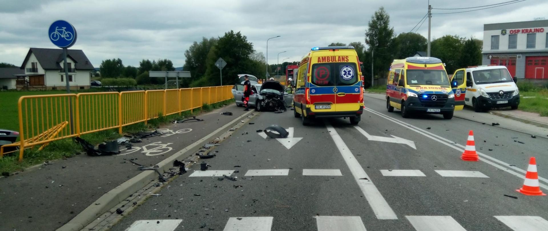 Zdjęcie przedstawia miejsce wypadku na drodze wojewódzkie. Na jezdni stoją 2 ambulanse, a w głębi widać jeden ze zniszczonych aut.