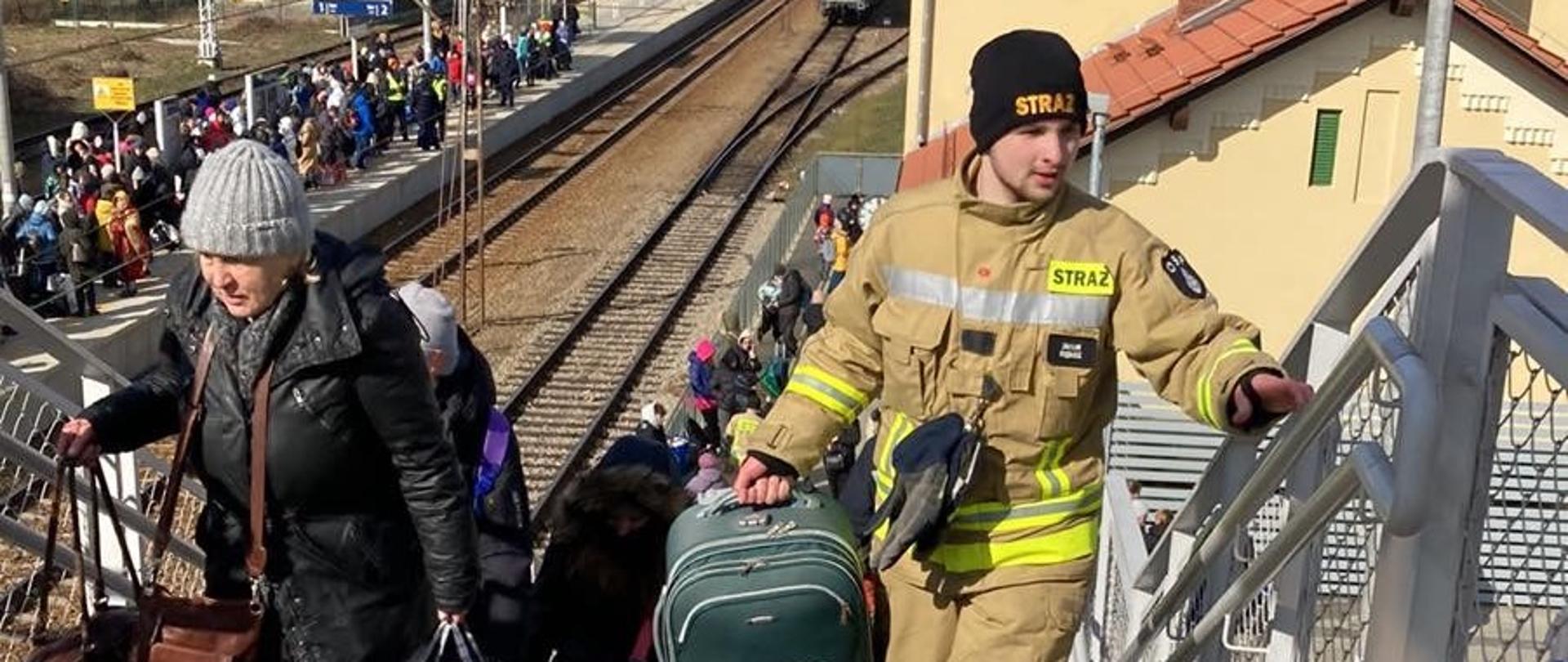 Strażak niesie walizkę i wchodzi po schodach, obok niego idzie kobieta niosąca podręczną torbę. W tle widoczny peron kolejowy na którym stoi duża ilość osób. 