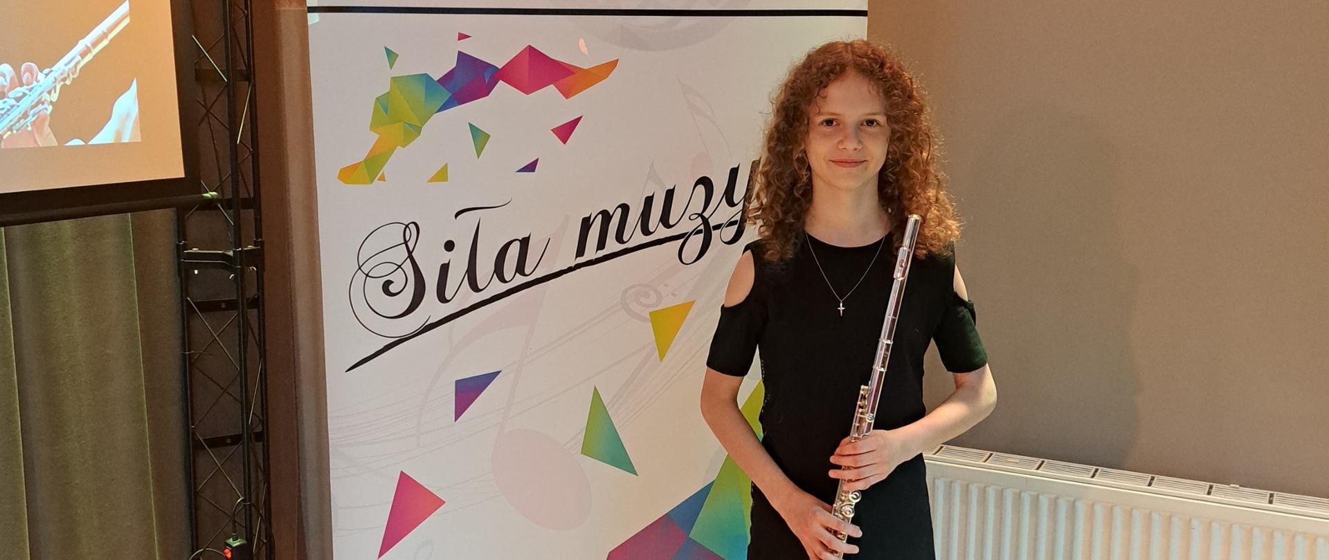 Na zdjęciu znajduje się dziewczynka ubrana w czarną sukienkę, trzymająca flet poprzeczny, w tle baner z napisem Siła muzyki i kolorowymi elementami na białym tle