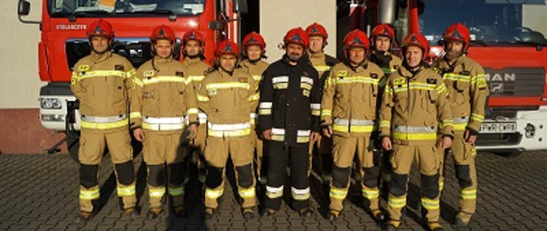 Odział strażaków stojący na tle dwóch wozów bojowych. Pośrodku w wyróżniającym się mundurze stoi odchodzący ze służby funkcjonariusz