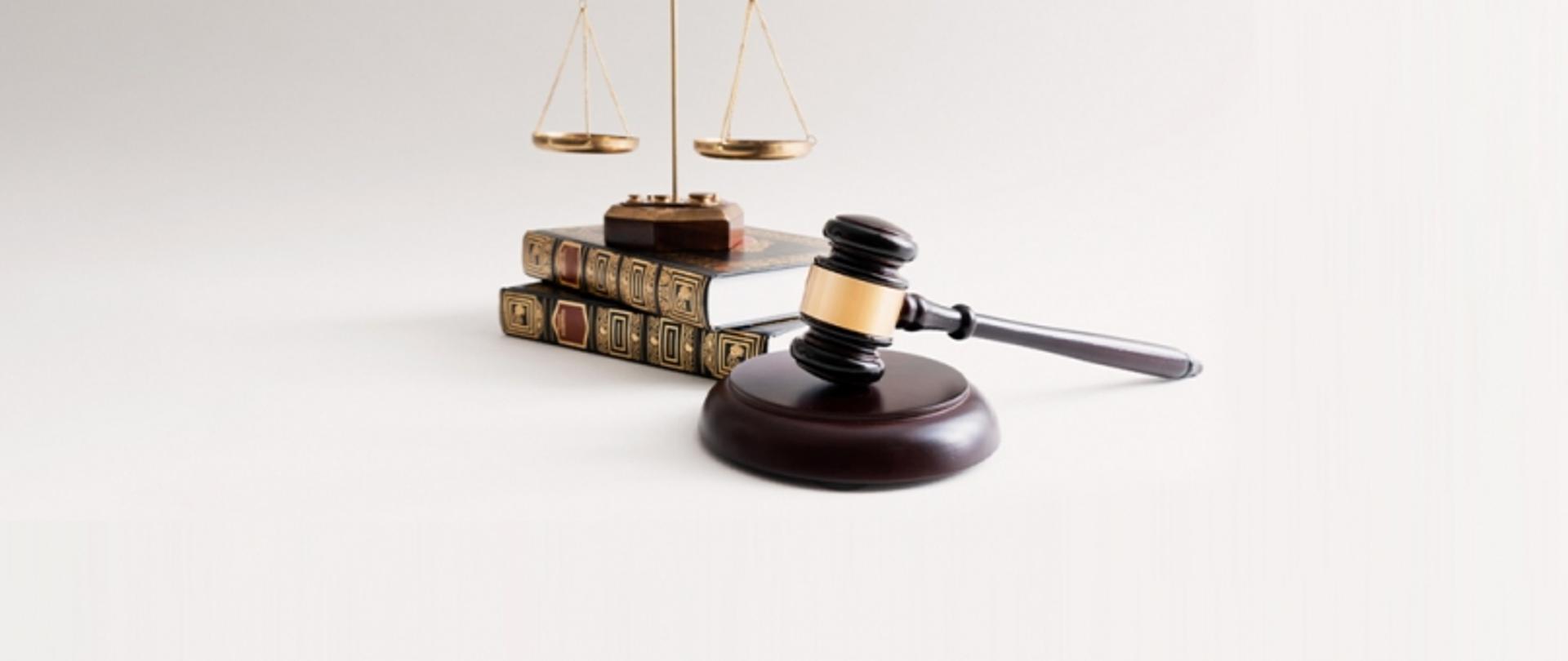 Młotek sędziego na podstawce, obok książki prawnicze i waga jako symbol prawa i sprawiedliwości.
