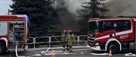 Strażacy prowadzą akcję gaśniczą przy domu jednorodzinnym, z którego unosi się gęsty dym; na drodze stoją dwa wozy strażackie, a węże pożarnicze są rozłożone na jezdni.