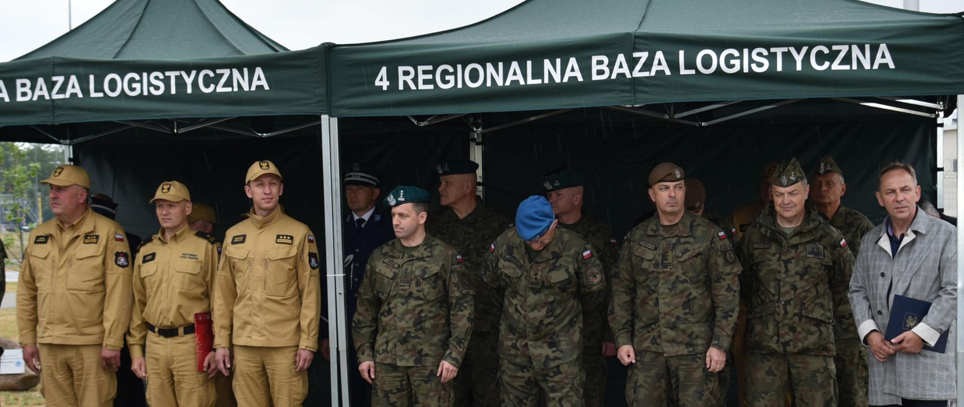 grupa strażaków oraz żołnierzy stoid pod zielonym namiotem na dużym placu