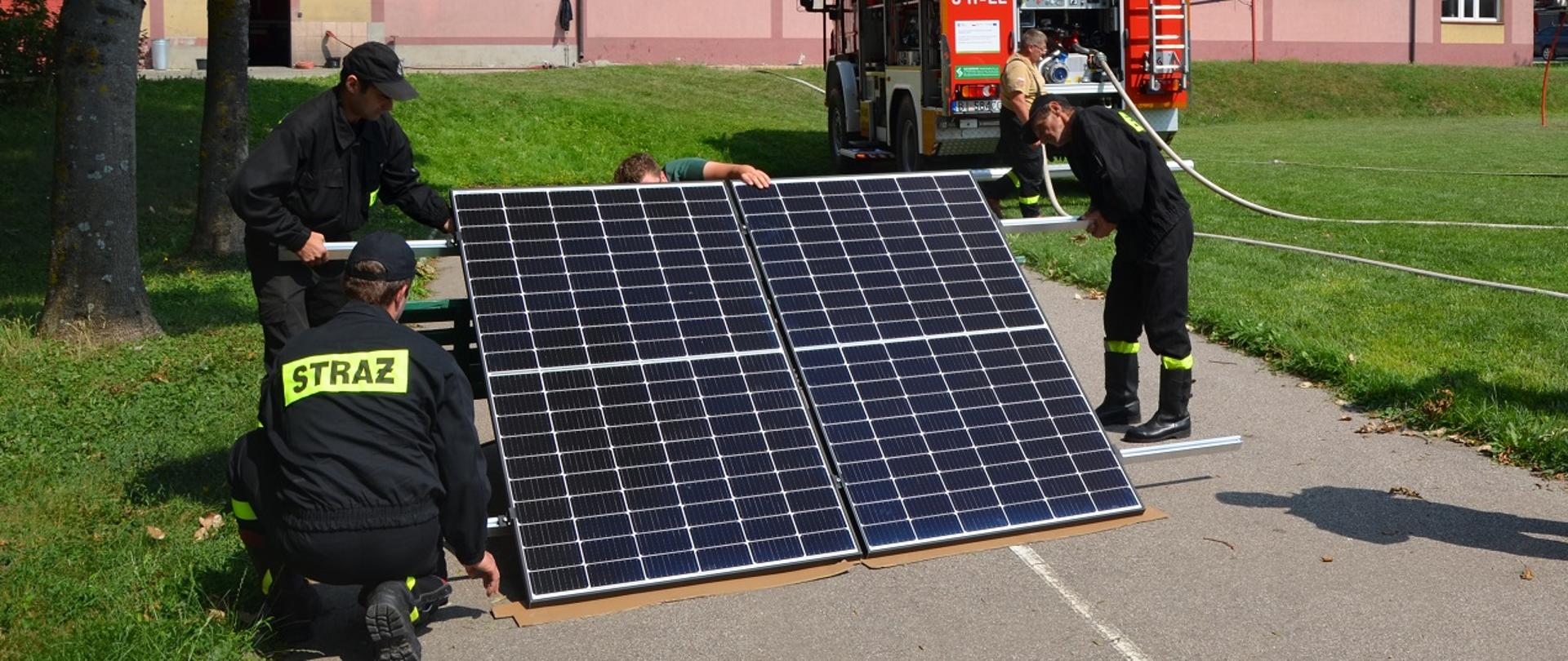 Zdjęcie obrazuje strażaków oglądających panele fotowoltaiczne w głębi samochód strażacki i budynek JRG nr 1. 