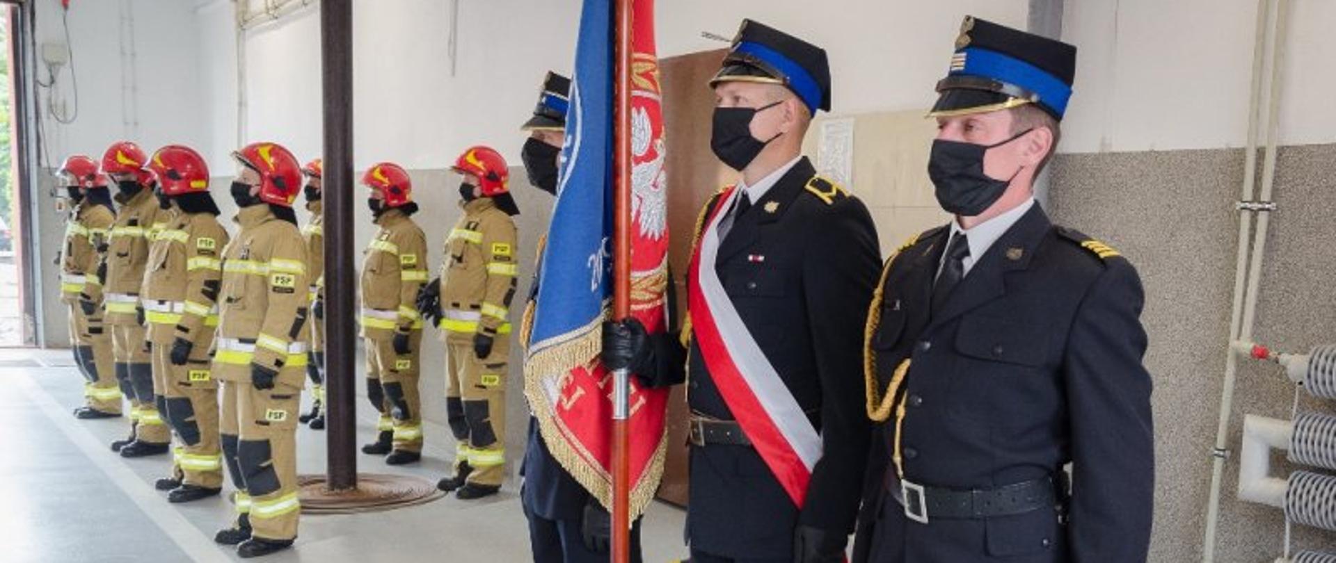 Samochody i odznaczenia dla mazowieckich strażaków – na zdjęciu poczet sztandarowy. Na drugim planie czterech strażaków w piaskowych mundurach i czerwonych hełmach. Zdjęcie zrobione w garażu jednostki.