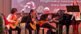Na zdjęciu przedstawiony jest występ instrumentalno-wokalny podczas szkolnego koncertu świątecznego. Na scenie siedzi trzech uczniów grających na gitarach klasycznych, a obok nich stoi uczennica grająca na flecie przy pulpicie z nutami. W tle widoczny jest fortepian oraz ekran z bożonarodzeniową grafiką i napisem „Mizerna Cicha, Gdy śliczna Panna” wraz z informacją o wykonawcach. Scena udekorowana jest świątecznie – widać choinkę, skrzata oraz drobne dekoracje. Na pierwszym planie znajduje się publiczność siedząca na widowni. Całość oddaje uroczysty i świąteczny charakter koncertu.