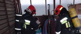 Na zdjęciu widać dwóch strażaków z aparatami powietrznymi na plecach którzy zabezpieczają wyciek gazu z butli znajdującej się w pomieszczeniu magazynowym na terenie zakładu.