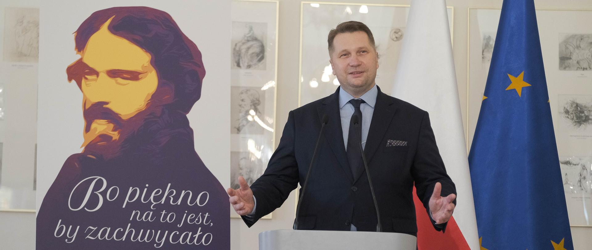 Wizyta Ministra Edukacji i Nauki w Liceum Ogólnokształcącym im. Cypriana K. Norwida w Radzyminie, minister Przemawia do mikrowonu podczas wuzyty. W tle z lewej strony plansza z portretem Norwida oraz napis bo piękno na to jest by zachwycało. Za ministrem w tle flaga Polski i Unii Europejskiej 