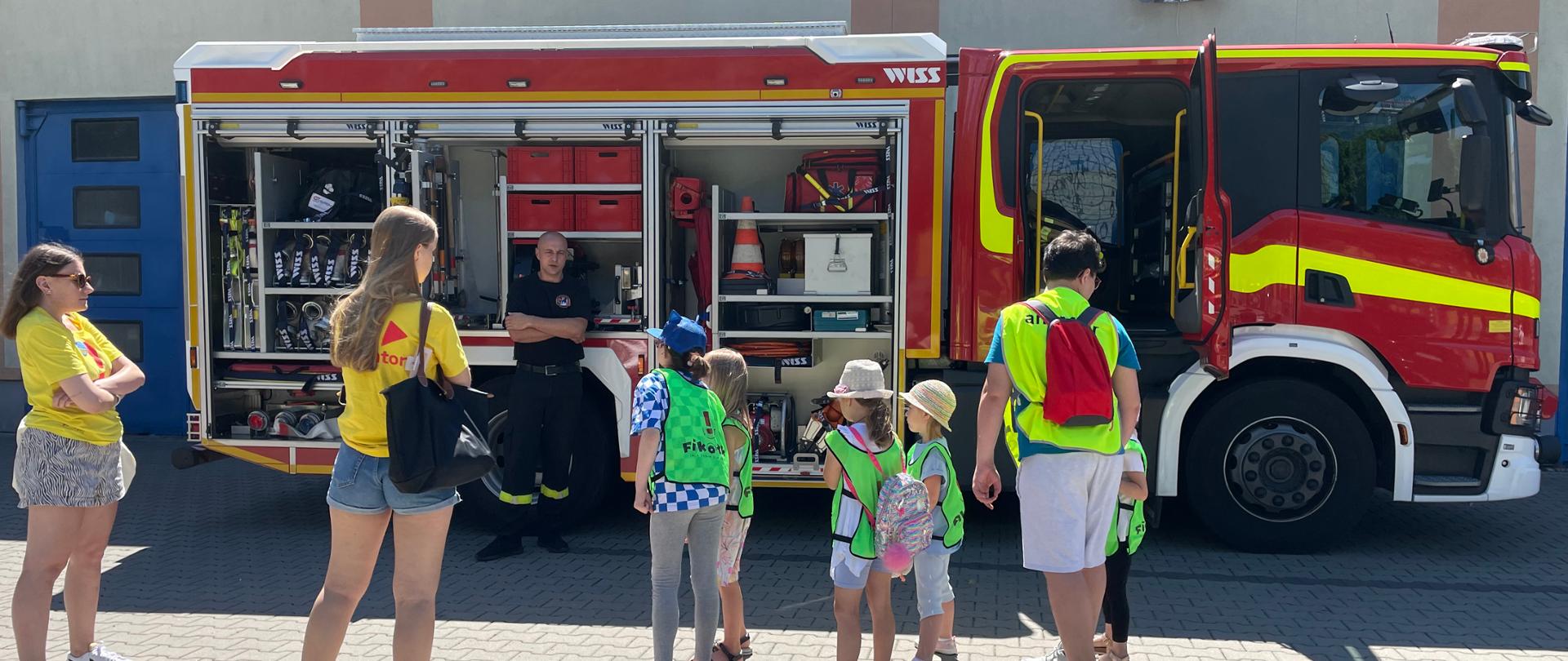 Na zdjęciu widoczny jest samochód strażacki, strażak pokazujący wyposażenie samochodu oraz grupa dzieci z opiekunami