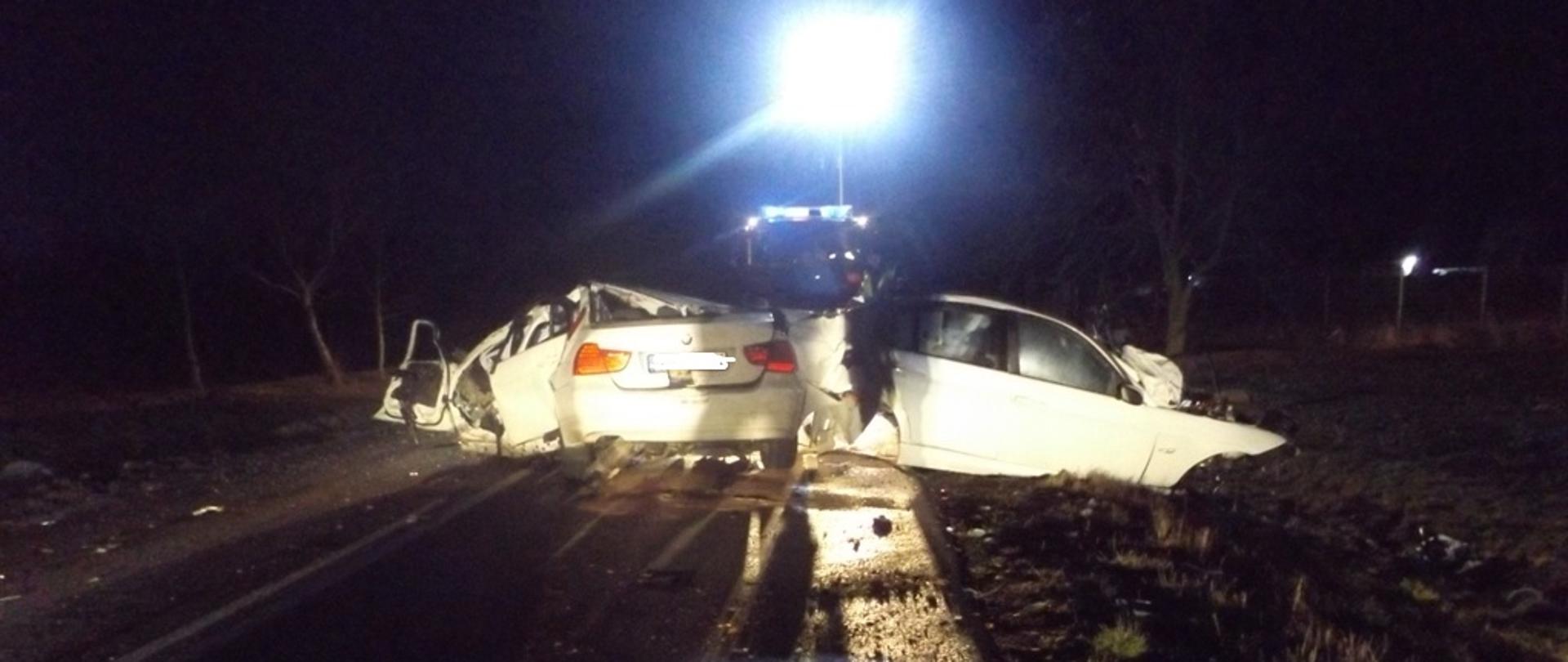 Samochód marki BMW został rozpołowiony po uderzeniu w drzewo. Części samochodu leżą rozrzucone w promieniu kilkunastu metrów. W tle widać samochód ratowniczo-gaśniczy oświetlający miejsce zdarzenia.
