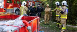 zdjęcie przedstawia Zastępcę Kujawsko-Pomorskiego Komendanta Wojewódzkiego PSP witającego się ze strażakami przy buforze wodnym. W tle samochody gaśnicze i zbiornik wodny. Widocznie na zdjęciu również strażacy w trakcie ćwiczeń 