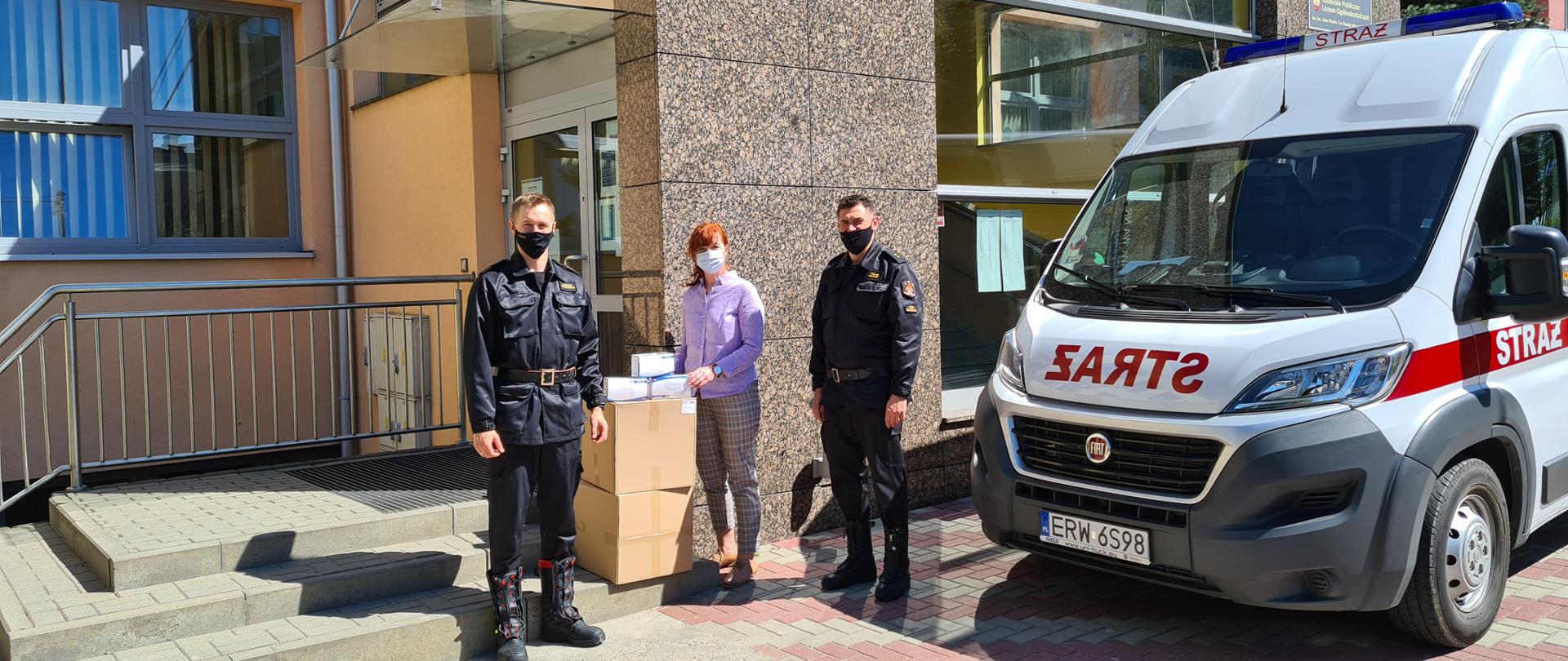 Zdjęcie przedstawia trzy osoby stojące przed wejściem do szkoły, a między nimi stoją 2 papierowe pudła z maseczkami. Na lewo stoi mężczyzna ubrany w strój strażacki, a na prawo kobieta ubrana w brązowe spodnie i jasny sweter oraz drugi mężczyzna ubrany w strój strażacki. Na prawo stoi również biało czerwony samochód FIAT Dukato. 