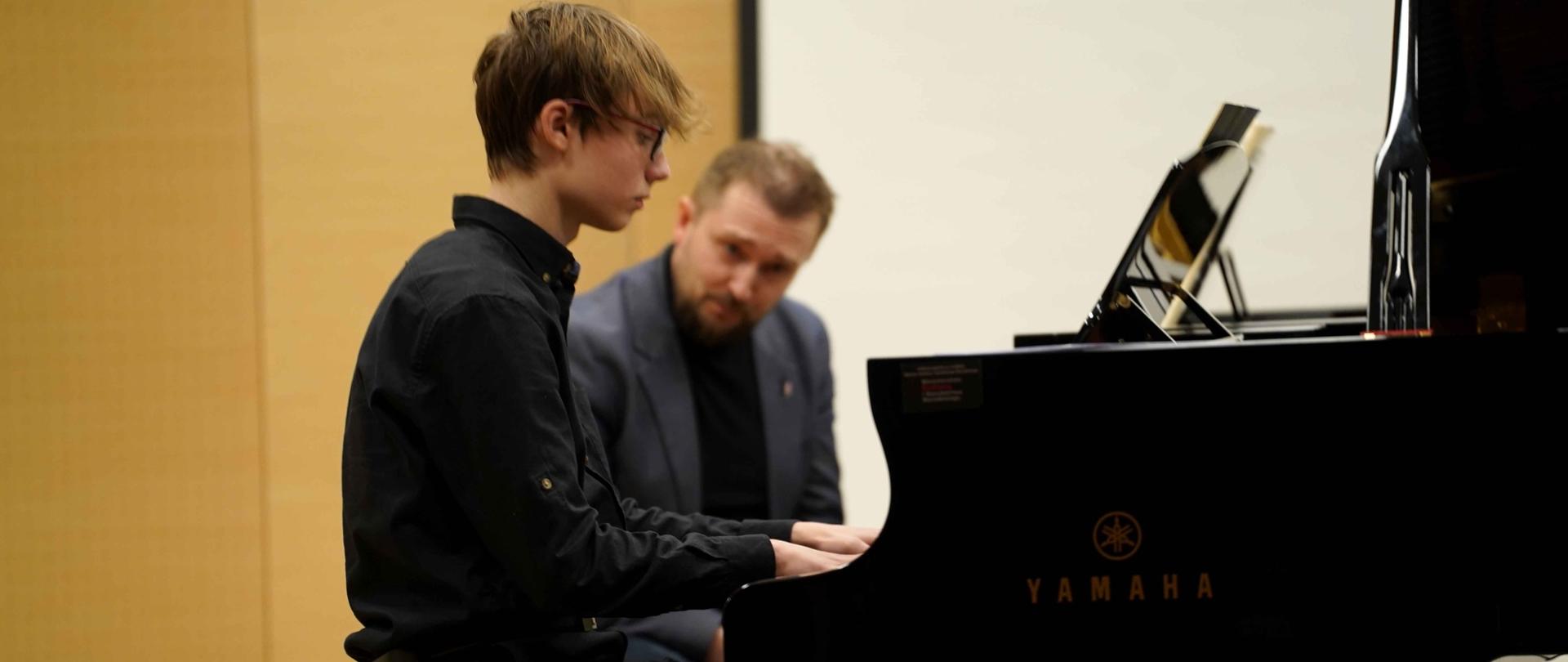 zdjęcie przedstawia ucznia grającego na jednym z dwóch fortepianów na scenie sali koncertowej, w tle prowadzący warsztaty przyglądający się w grze ucznia